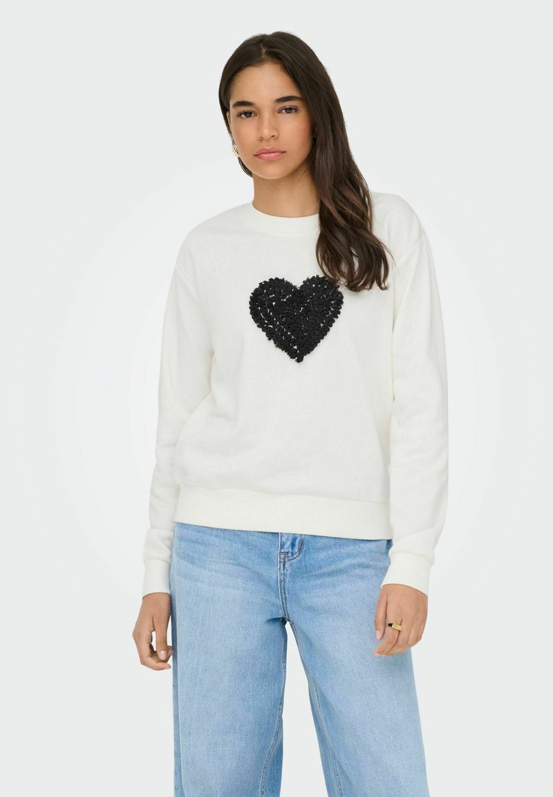 Sweat-shirt blanc avec un cœur texturé en sequins noirs sur la poitrine. Présente un col rond et des poignets côtelés, associé à un jean bleu clair.