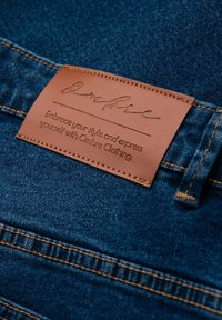 Jeans de mezclilla de tela azul oscuro, con una parche de cuero marrón con texto en relieve, bordes cosidos y hilo naranja en contraste.