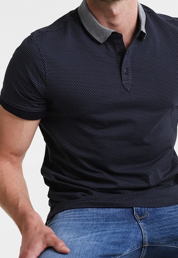 Homme portant un polo bleu marine à manches courtes avec petits points blancs et un col gris, associé à un jean bleu.