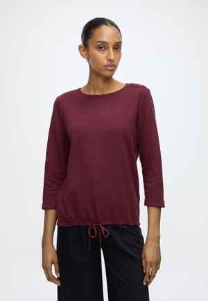Femme portant un haut à manches longues bordeaux avec des détails de boutons à l'épaule et un cordon de serrage à l'ourlet, associée à un pantalon noir, debout sur un fond blanc.