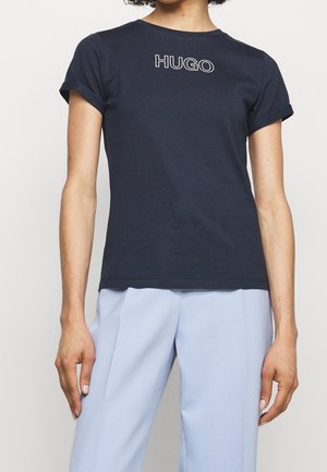 Osoba ubrana w granatowy T-shirt z logo "HUGO" na piersi oraz jasnoniebieskie spodnie, stojąca na jednolitym białym tle.