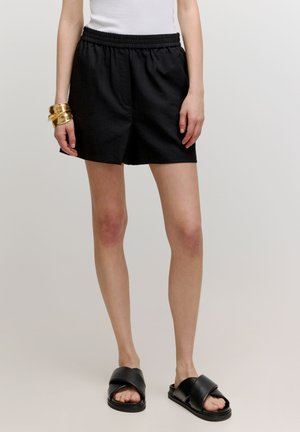 Shorts en lin noir avec une taille élastique, poches latérales, coupe décontractée, associés à des sandales noires à talon plat sur pieds nus.