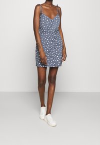 Nainen, joka on pukeutunut siniseen olkaimettomaan mini mekkoon, jossa on valkoisia täpliä, napit yksityiskohtina ja solmittavat olkaimet, sekä valkoisiin lenkkareihin, seisoo yksinkertaisen taustan edessä.