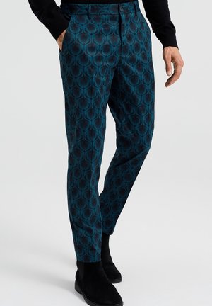 Mężczyzna w dopasowanych spodniach w kolorze morskim z wzorem, czarnej bluzce z długim rękawem oraz czarnych sznurowanych botkach zamszowych, z jedną ręką w kieszeni.