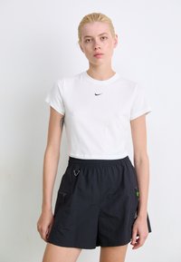 Balta trumpa marškinėliai su juodu Nike logotipu, poruoti su juodais lengvais šortais, turinčiais šonines kišenes ir raištelių liemenį.