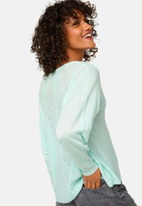 Femme aux cheveux bouclés portant un pull bleu clair sourit, posant avec une main dans la poche arrière d'un jean gris foncé.