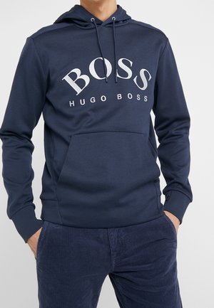 Uomo che indossa una felpa con cappuccio Hugo Boss blu navy con coulisse e tasca frontale, abbinata a pantaloni di velluto a costine blu navy.