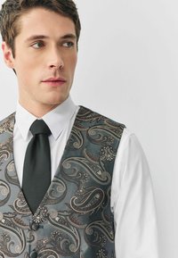 Ciemnozielona kamizelka w wzór paisley wykonana z teksturowanej tkaniny, z dodatkami w kolorze srebrnym oraz dopasowaną czarną muszką na białej koszuli.