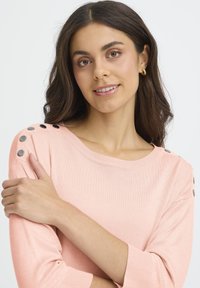 Pull rose clair à texture côtelée, avec des boutons métalliques noirs sur les épaules et un col rond, style manches trois-quarts.