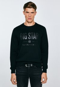 Junger Mann, der einen schwarzen Pullover mit dem Aufdruck "BIG STAR Your Life. Your Jeans." und schwarze Jeans trägt, steht vor einem einfachen, hellen Hintergrund.