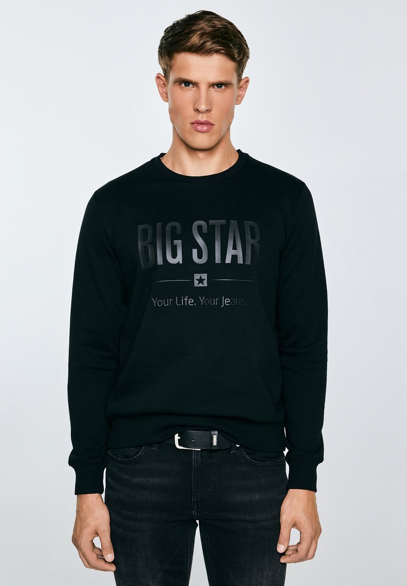 Junger Mann, der einen schwarzen Pullover mit dem Aufdruck "BIG STAR Your Life. Your Jeans." und schwarze Jeans trägt, steht vor einem einfachen, hellen Hintergrund.