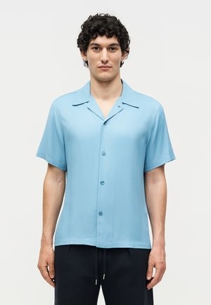 Νέος άνδρας με σγουρά μαλλιά, φορώντας ένα ανοιχτό μπλε κοντομάνικο πουκάμισο με κουμπιά και μαύρο παντελόνι με κορδόνια σε μονόχρωμο φόντο.