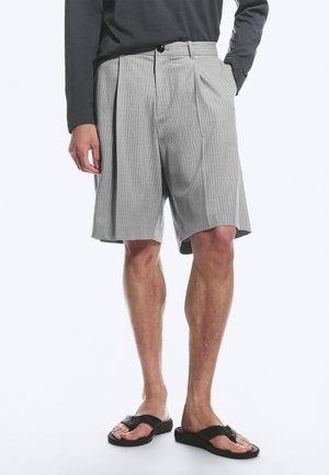 Mand iført grå ternede plisserede shorts, sort langærmet top og sorte sandaler, stående med den ene hånd i lommen.