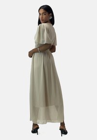 Maxi dress in light beige chiffon with short puff sleeves and a smocked waist, featuring a flowing skirt and subtle sheen. Black heeled sandals.