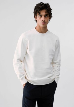 Jeune homme portant un sweat-shirt blanc uni à manches longues et un pantalon foncé, debout les mains dans les poches sur un fond clair.