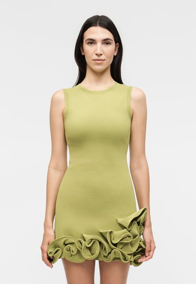 Femme aux longs cheveux foncés portant une robe vert clair sans manches, cintrée, avec des volants le long de l'ourlet inférieur.