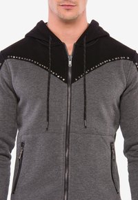 Graue Kapuzenjacke mit einem schwarzen Schultereinsatz, verziert mit silbernen Nieten entlang der oberen Naht und zwei seitlichen Reißverschlusstaschen.