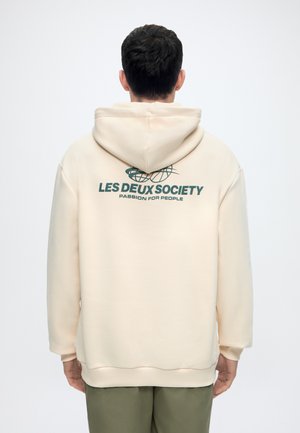 Homme de dos portant un sweat à capuche crème avec le texte « LES DEUX SOCIETY PASSION FOR PEOPLE » et un graphisme de globe au dos, associé à un pantalon olive.