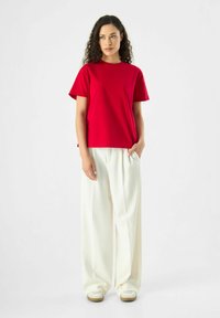 Rotes Baumwoll-T-Shirt mit kurzen Ärmeln, lockere Passform, kombiniert mit weißen weitgeschnittenen Hosen. Das Model steht mit einer Hand in der Tasche.