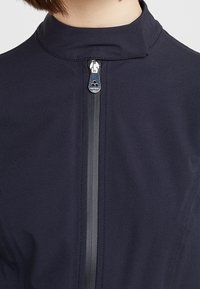 Navy zip-up garment with a high collar, featuring a silver zipper and a vertical black stripe. Smooth fabric texture with subtle stitching details.