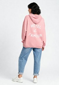 Felpa rosa con cappuccio con "HÔTEL de l’amour" stampato in bianco sulla schiena, abbinata a jeans azzurri con risvolto e sneakers bianche.