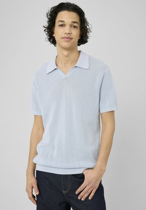 Jeune homme aux cheveux bouclés portant un polo tricoté bleu clair à manches courtes et un jean foncé, debout devant un fond gris uni.