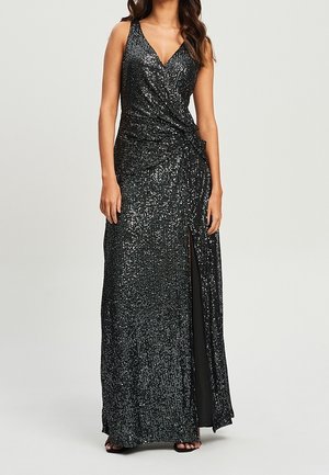 Ärmelloses schwarzes Paillettenkleid mit V-Ausschnitt, Seitenschlitz und gerafftem Taillendetail, kombiniert mit schwarzen offenen High-Heels.