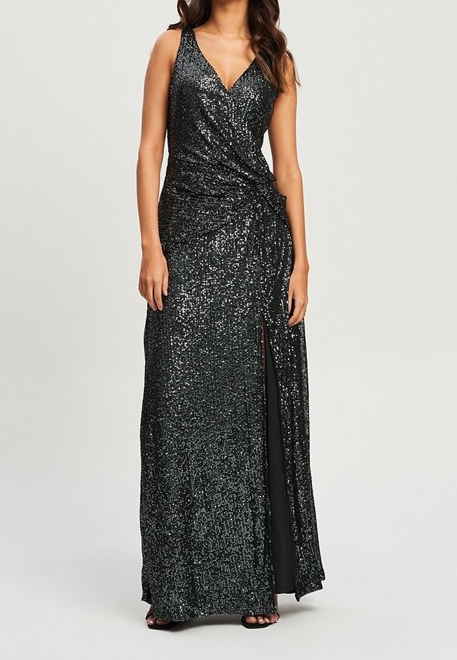 Robe noire sans manches ornée de sequins, décolleté en V, fente latérale et détail froncé à la taille, portée avec des talons ouverts noirs.