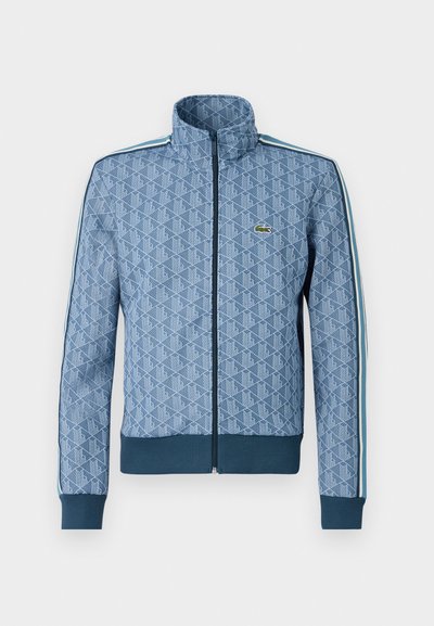 Veste zippée bleue avec un motif géométrique, des poignets et un ourlet côtelés, et des bandes latérales contrastantes. Présente un petit logo sur la poitrine.