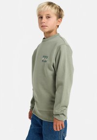 Sweatshirt verde com decote redondo, punhos e barra canelados, com um pequeno patch de design de onda no lado esquerdo do peito. Ajuste casual.