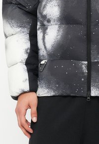 Jachetă puf cu design în degradé alb-negru, având închidere cu fermoar, suprafață texturată și un mic carabină argintie pe lateral.