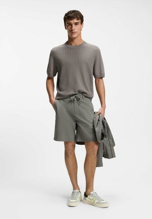 Ung mand stående iført en grå ribstrikket kortærmet sweater, grå shorts, holder en grå jakke og hvide sneakers med lysegrønne detaljer.