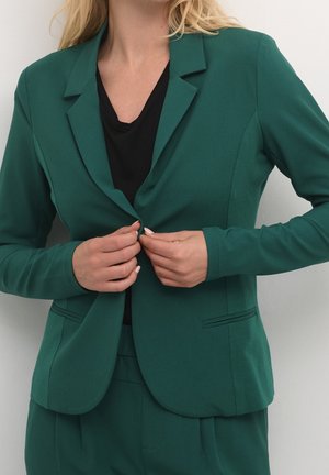 Femme boutonnant un blazer vert foncé sur un haut noir, montrée du cou aux hanches sur un fond uni.