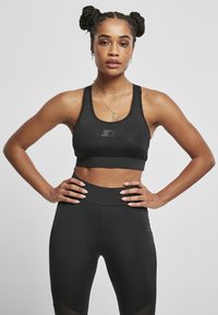 Čierny športový crop top s logom, s lodičkovým výstrihom a širokými ramienkami. V kombinácii s čiernymi legínami s vysokým pásom a hladkým povrchom.