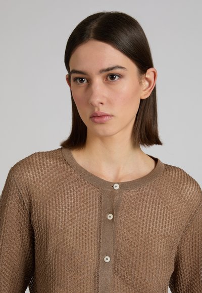 Jonge vrouw met recht, donkerbruin haar, gekleed in een bruin gebreid vest met knopen, kijkt lichtjes opzij.