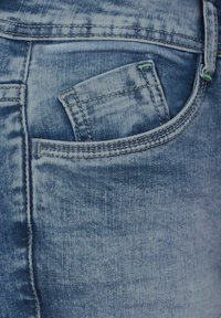 Jean en denim bleu avec une texture usée et des coutures visibles, présentant une petite poche avant avec des accents de couture verte.