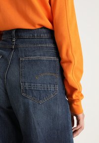 Ciemnoniebieskie dżinsy z tylną kieszenią wykończoną zakrzywionym pomarańczowym szwem, w połączeniu z pomarańczową bluzką z długim rękawem. Widoczna fakturowana tkanina.