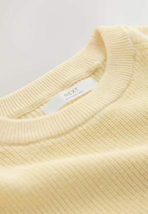 Žuti pulover s rebrastim uzorkom, okruglim izrezom i mekanom teksturom. Na etiketi piše "NEXT, made in Bangladesh."
