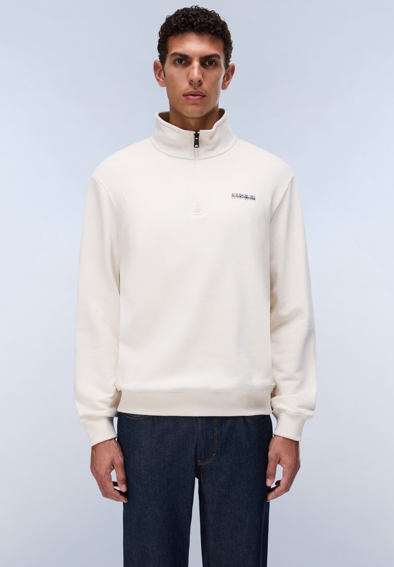 Homme aux cheveux courts et bouclés portant un pull crème à fermeture éclair quart de zip et un jean bleu foncé, debout devant un fond clair uni.