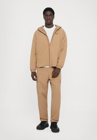 Veste zippée marron avec capuche, poignets élastiques et poches, associée à un pantalon fuselé assorti. Portée avec un t-shirt blanc à col rond et des chaussures noires.