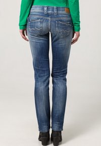 Jeans de denim azul ajustados con detalles de lavado claro, dos bolsillos traseros con costuras curvas y un parche de cuero marrón en la cinturilla.
