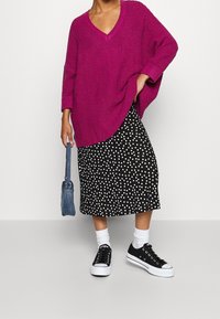 Pull en maille magenta à col en V, associé à une jupe noire à pois blancs. Des baskets noires et un sac à main en jean complètent la tenue.