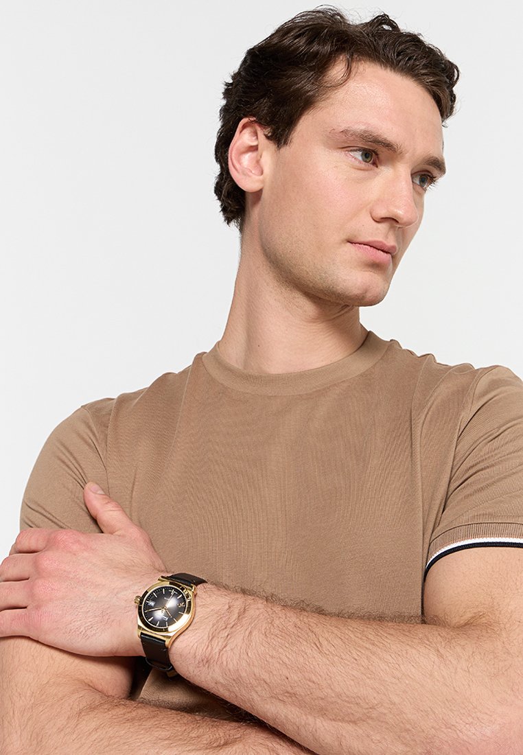 T-shirt marron à rayures avec des accents noir et blanc, associé à une montre en or et noir avec un cadran rond et un bracelet en cuir.