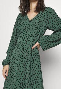 Grünes Kleid mit V-Ausschnitt, langen Ärmeln und schwarzen Punkten. Hergestellt aus einem weichen Material mit lockerer Passform und seitlichen Taschen.