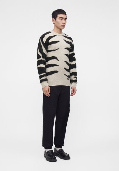 Homme debout portant un pull beige avec des rayures tigre noires, un pantalon noir, des chaussettes blanches et des mocassins noirs sur un fond clair uni.