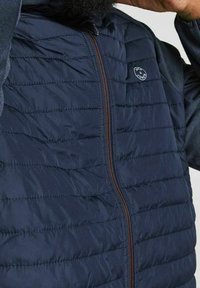 Mörkblå puffjacka med horisontella quiltade mönster, en bronze dragkedja och en logotypapplikation på vänster bröstområde. Strukturerad tyg.