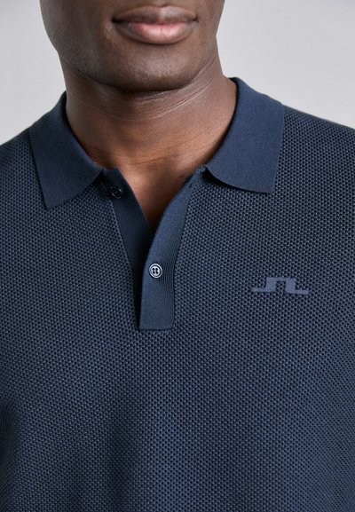 Camisa polo azul marino con un tejido texturizado, presenta un cuello clásico, una tapeta de dos botones y un pequeño logo en el pecho.