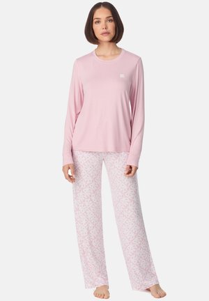 Femme debout pieds nus portant un haut à manches longues rose clair et un pantalon blanc avec des motifs géométriques roses sur un fond blanc.