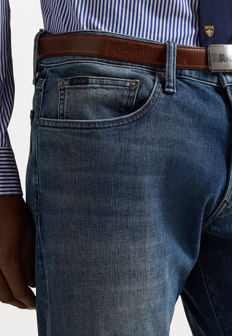 Denim jeans in a medium blue hue with a slight fade, featuring a patch label on the pocket, paired with a brown leather belt.