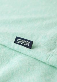 Tessuto verde menta chiaro con una texture morbida, dotato di un'etichetta blu scuro con la scritta "SUPERDRY" in lettere bianche.